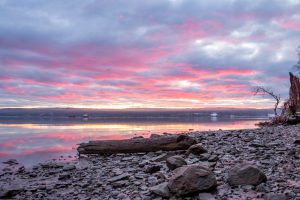 A photo of rocks and a log on the shoreline of the Hudson River during a January sunrise.
