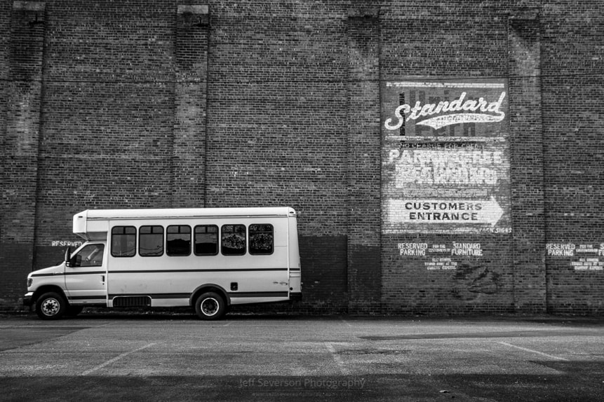 A black and white photo of a parking lot with old signage painted on a brick building with a bus parked in front of it.