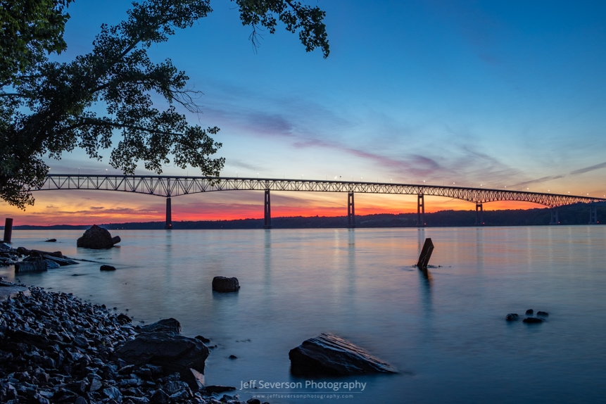 A photo of the first stages of a June sunrise appearing behind the Kingston-Rhinecliff Bridge as seen from the shores of the Hudson River at Charles Rider Park.