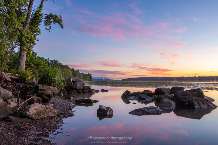 A photo of a June sunrise from along the shores of the Hudson River with clouds reflected in the water.