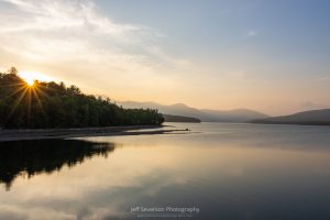 A photo of the sun setting behind the treeline with Ashokan Reservoir in the foreground and the Catskill Mountains in the background.