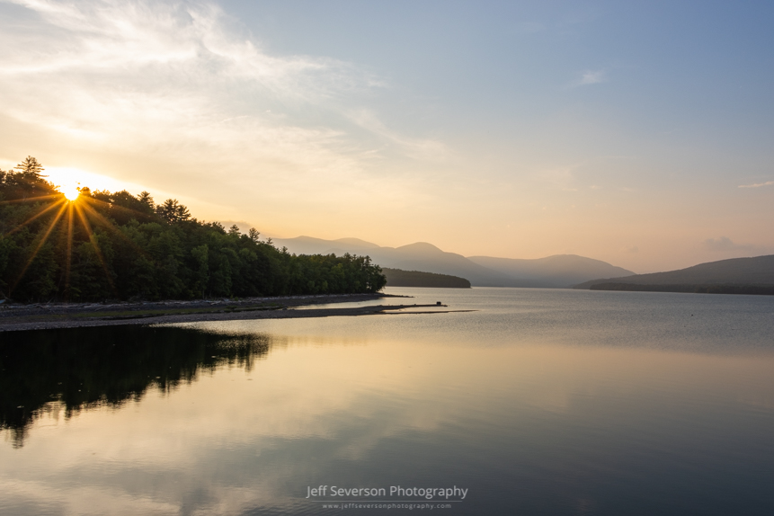 A photo of the sun setting behind the treeline with Ashokan Reservoir in the foreground and the Catskill Mountains in the background.