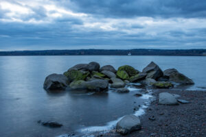 A long exposure photograph of the shoreline of the Hudson River at Lighthouse Park during the end of the blue hour before sunrise.