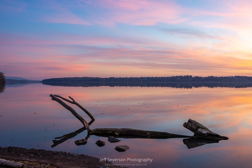 A photo of driftwood silhouetted in the water during a sunrise while looking North up the Hudson River in mid-April from the shore of Lighthouse Park in Ulster Park.