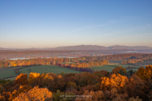 A landscape view of the fall foliage in Northern Dutchess County, with the Kingston-Rhinecliff Bridge and the Catskill Mountains in the distance, from atop the Ferncliff Fire Tower during the golden hour of sunrise on an October morning.